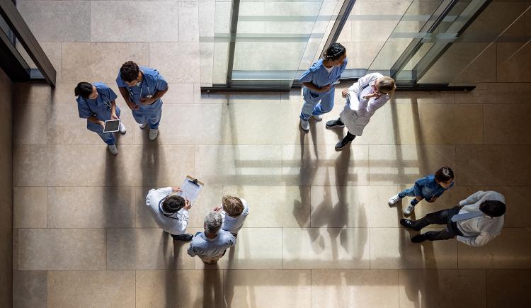 An overhead view of nine people in the entryway of a hospital, standing in groups of two or three, many dressed in medical gear or lab coats, carrying clipboards and digital devices.
