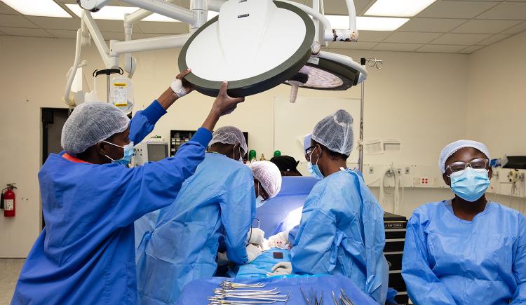 A surgical team of doctors and nurses wearing blue scrubs, gauzy head coverings, and medical masks adjust lights, provide anesthesia, and perform other actions necessary for an operation, all gathered around the patient on the operating table. Health equity requires access to surgical care.