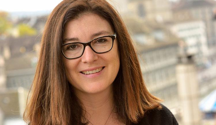 Portrait photo of a smiling woman with glasses and shoulder-length hair, sitting on a wall with a city skyline out of focus in the background.