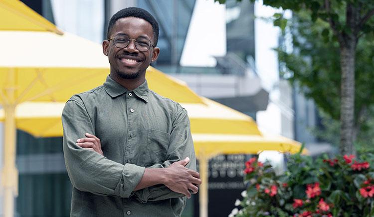 A smiling young Black man with glasses stands with arms crossed in front of picnic tables on the HMS Quad