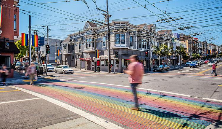 People in colorful outfits cross the street at an intersection with rainbow crosswalks in San Francisco’s Castro district.