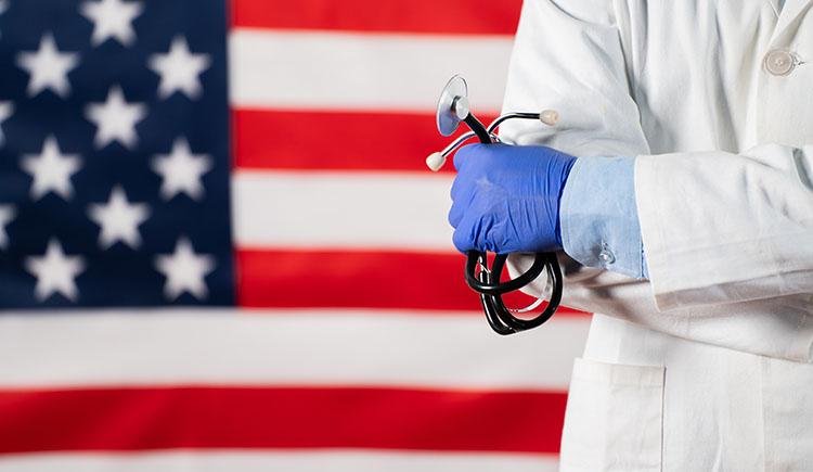 A health care practitioner wearing a white coat and holding a stethoscope stands in front of an American flag