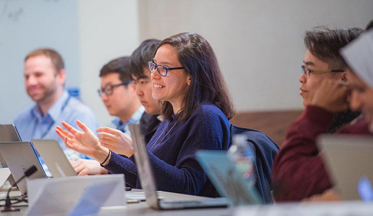 Six trainees at a conference table with laptops in front of them. The one at center speaks with a smile and gesturing hands.