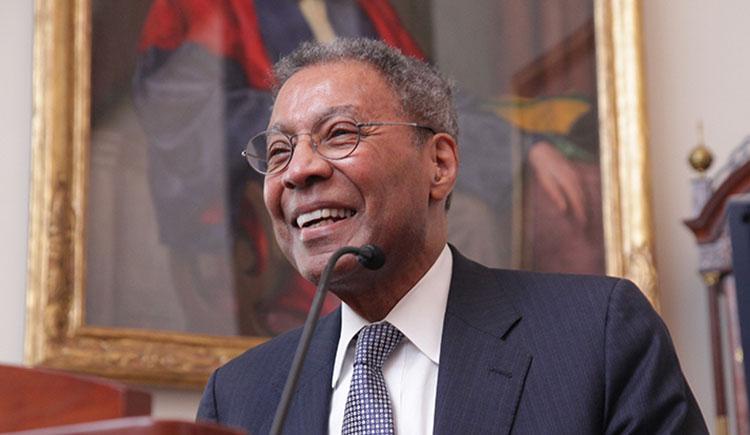 A man in a suit smiles at a lectern in front of a painting of someone in academic robes