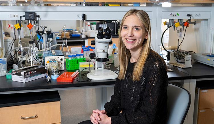 A researcher sits on a chair next to a microscope in a lab space