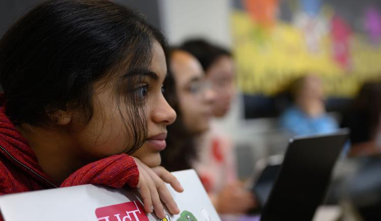 Focus on one student leaning on laptop with an HST sticker on the cover with several students in blurred background.