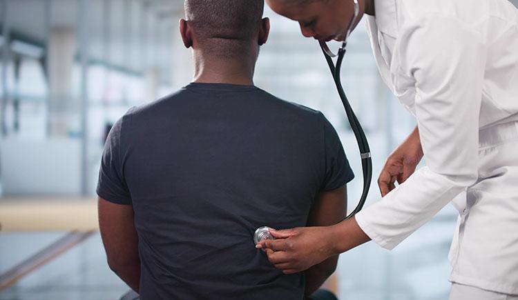 A doctor in a white lab coat uses a stethoscope to listen to the back of a seated patient wearing a dark t-shirt, in a bright, modern medical office.