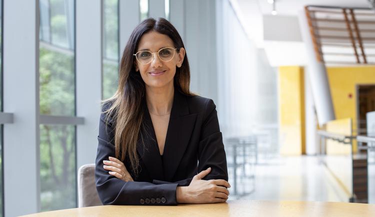 A woman with long, straight hair and glasses sits at a round wooden table in a modern, sunlit building with large windows. She is wearing a black blazer and smiling confidently with arms crossed.