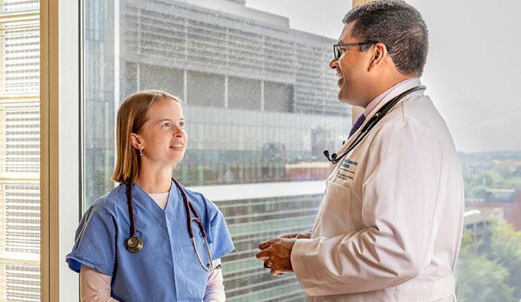 A woman in blue scrubs and a man in a white coat, both wearing stethoscopes, stand talking to each other in front of a window.