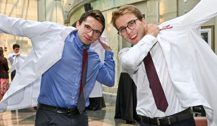 Two men put on white coats in a sun-filled atrium.
