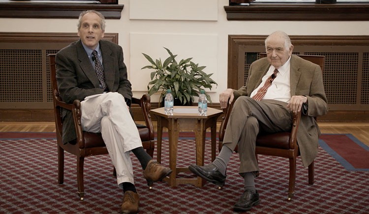 Two men in sports coats and ties sit in armchairs in front of a small table.