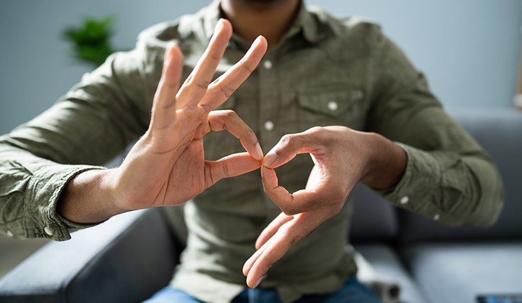 Close-up of two hands belonging to a person of color as they form the ASL sign for “interpreter.” The person’s head is unseen as they sit in a chair in a button-down shirt.