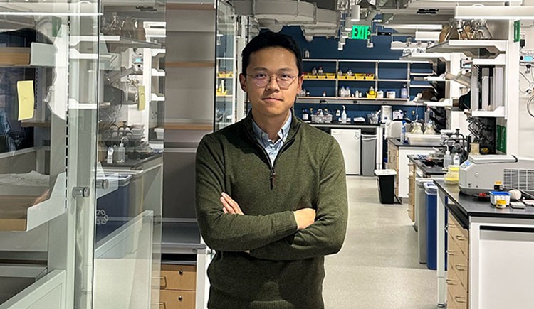 A man stands in a chemistry lab, with machinery on lab benches and reflections on glass partitions. 