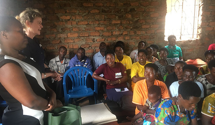 A group of adults seated closely together inside a simple brick-walled room listen attentively as two women stand at the front speaking. The audience includes men and women of different ages, some holding notebooks or bags, creating the feel of a community meeting or training session.