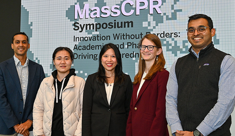 Five people in business clothes pose for a photo in front of a screen with the MassCPR symposium name