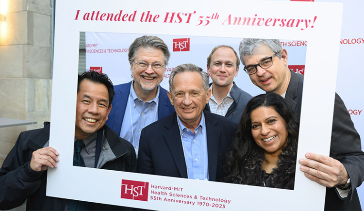 Six people pose, smiling, inside a large portable photo frame that says “Harvard-MIT Health Sciences & Technology 55th Anniversary 1970-2025”