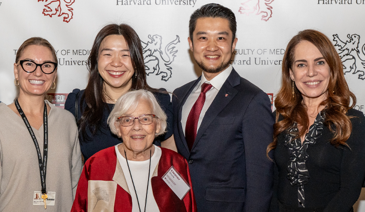 A group of adults stands in front of a backdrop displaying the Harvard University Faculty of Medicine logo. They are smiling and posing for a group photo. The group includes an older woman in a red and beige jacket standing in front. Everyone is dressed in professional or semiformal attire.