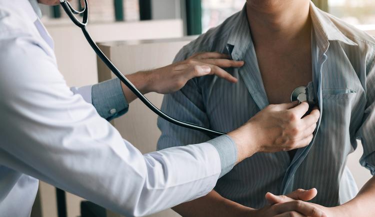 A person in a white jacket is using a stethoscope to listen to the heartbeat of person with a loosely buttoned striped shirt.