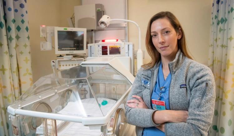 A woman in scrubs and a fleece stands with arms crossed next to a neonatal cradle.