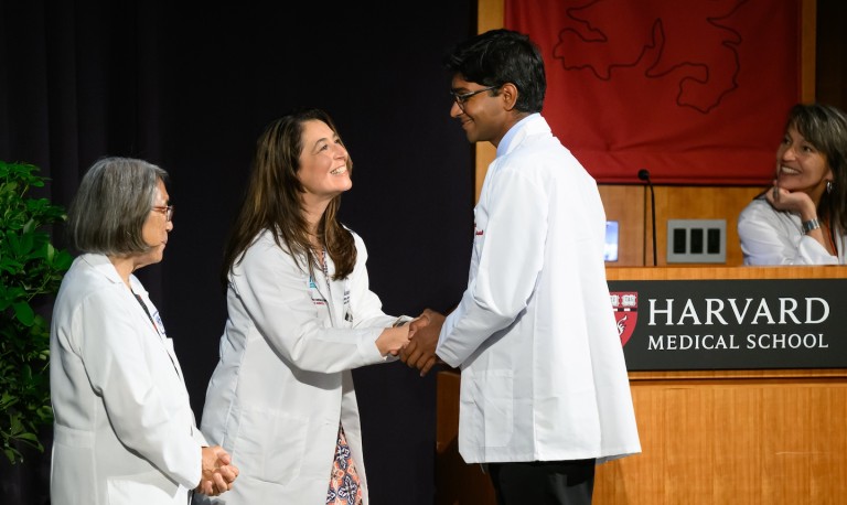 A new MD student receives a white coat while being greeted by three faculty from the academic society.