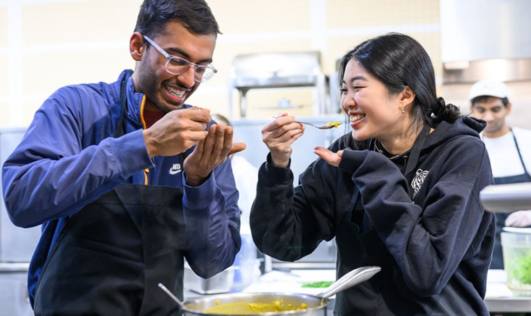 Two young adults wearing aprons taste a spoonful of food from a large pot.