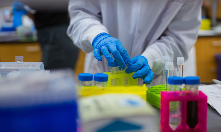 : Scientist wearing a white coat and blue gloves working in a lab setting.