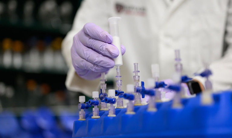 A researcher wearing purple gloves handles a syringe connected to laboratory equipment with multiple valves and tubes arranged in a blue rack