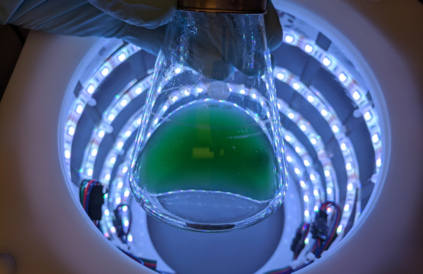 A flask of green liquid held by a gloved hand is silhouetted at the top of a cylinder with rings of lights