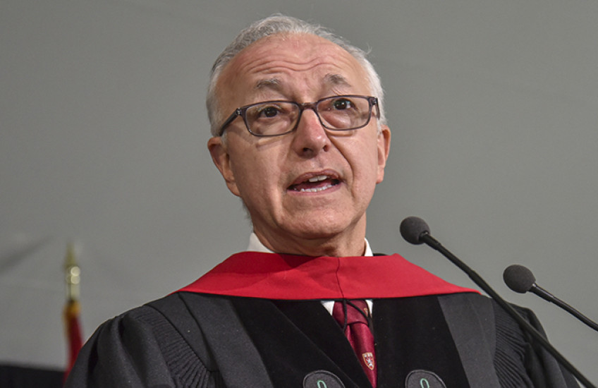 A gray-haired man with glasses and doctoral robes speaks into a microphone