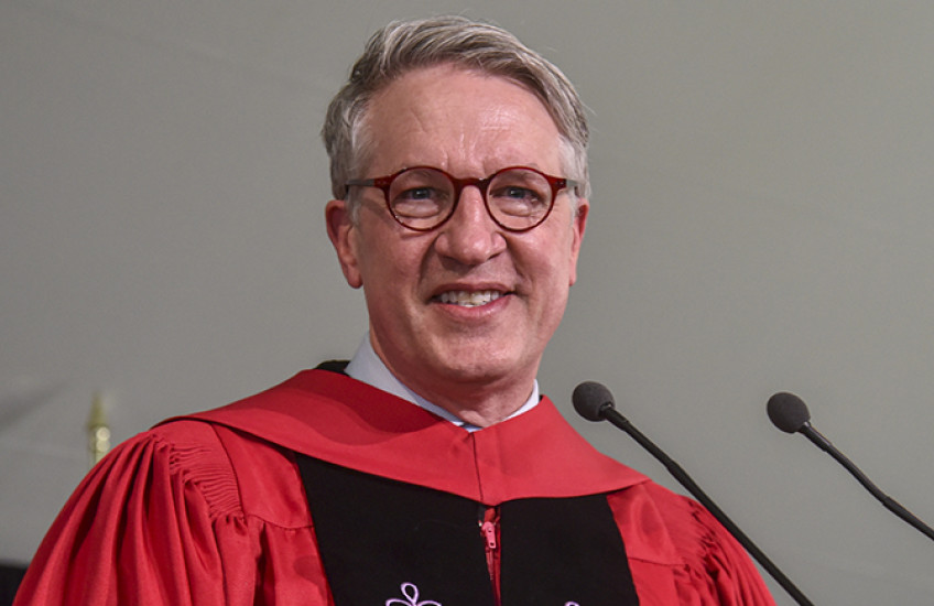A white man in glasses and red robes smiles at a microphone