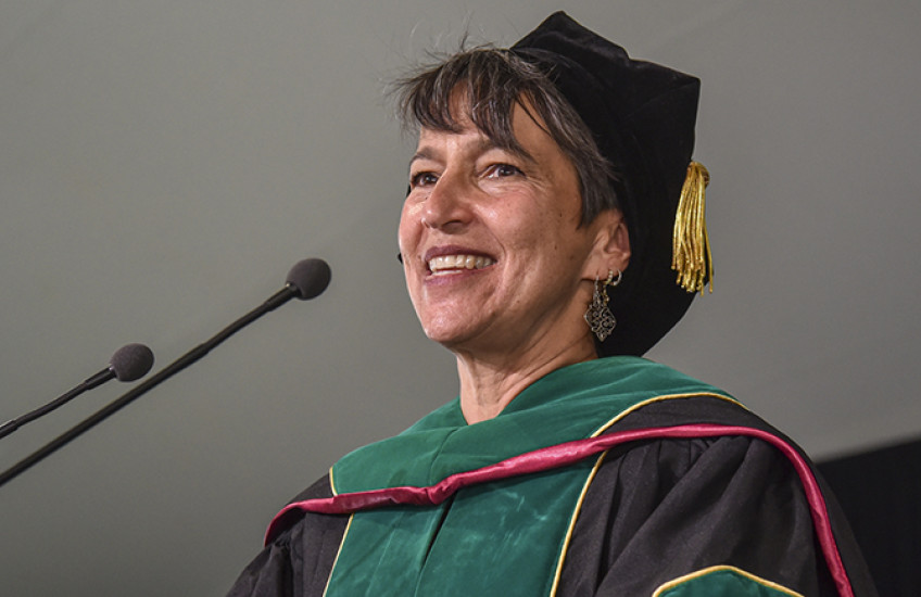 Close-up of a white woman with gray hair in doctoral robes smiling as she speaks into a microphone