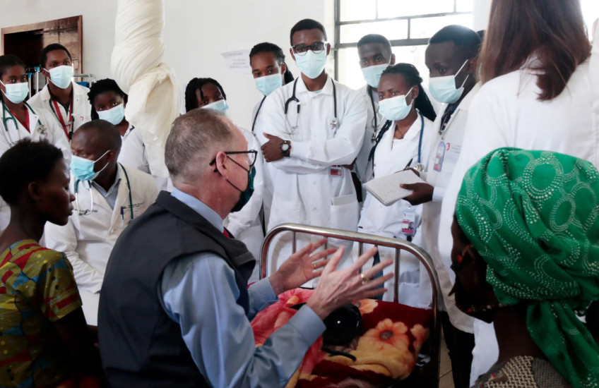 A line of women and men wearing white lab coats, medical masks, and stethoscopes stand facing three people, including Paul Farmer, sitting on hospital beds. 