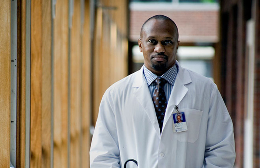 Stultz in a white coat with stethoscopes and hands in his pockets standing in a hallway