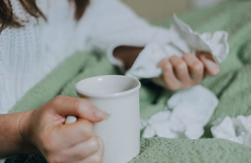 a person sitting in bed under a blanket, holding tea and surrounded by crumpled facial tissues