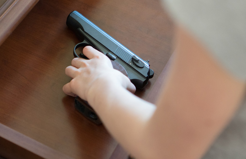 A child's hand reaches into an open drawer in a wooden dresser to pick up a black handgun with a brown handle.