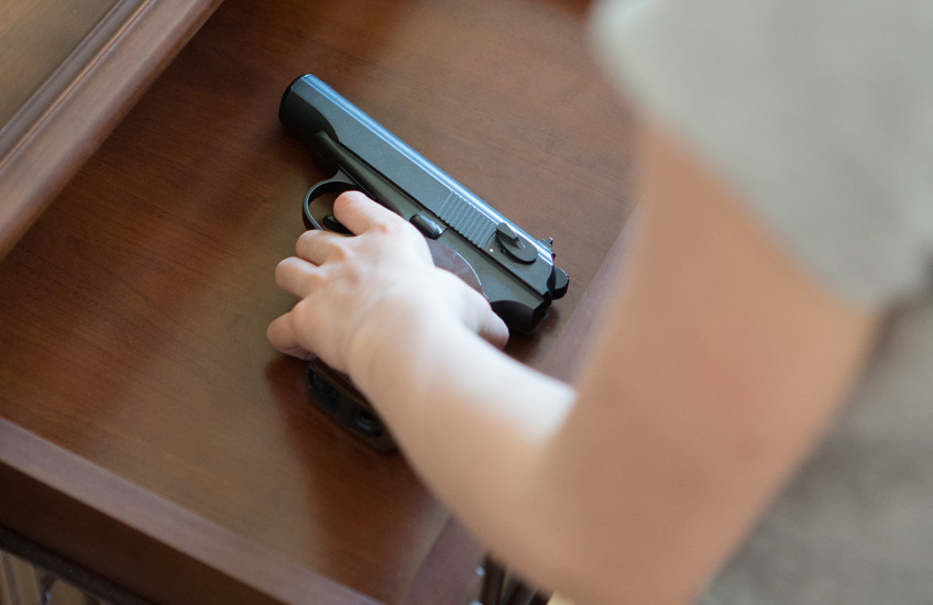 A child's hand reaches into an open drawer in a wooden dresser to pick up a black handgun with a brown handle.