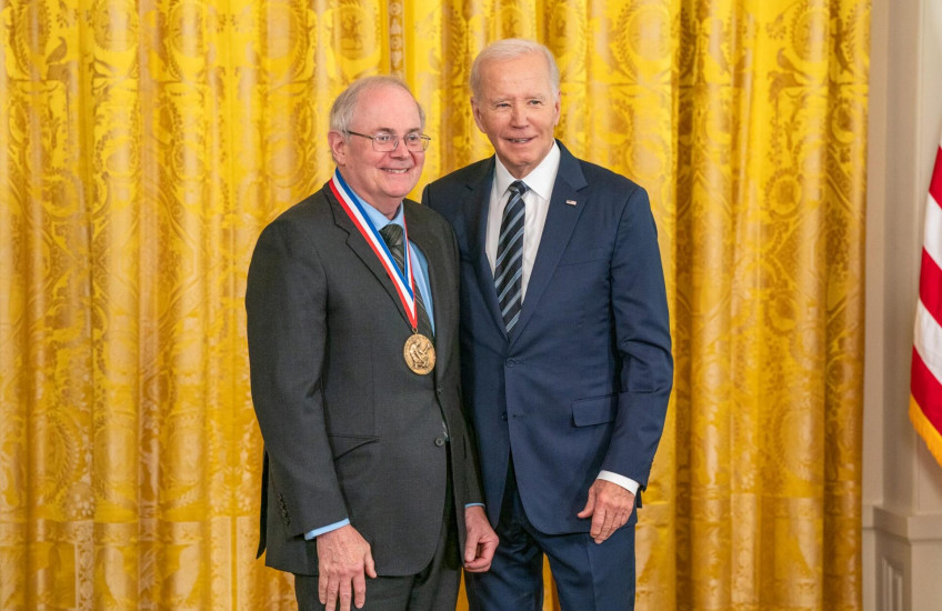 U.S. President Joseph Biden stands in a suit beside Gregory Petsko, a gray-haired white man also in a suit. Petsko wears his new National Medal of Science. Both are smiling. They stand before a gold curtain.