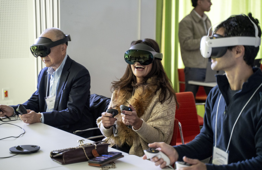 Two men and a woman sit at a conference table wearing large goggles that cover half their heads. The woman, sitting in the middle, laughs, showing white teeth.