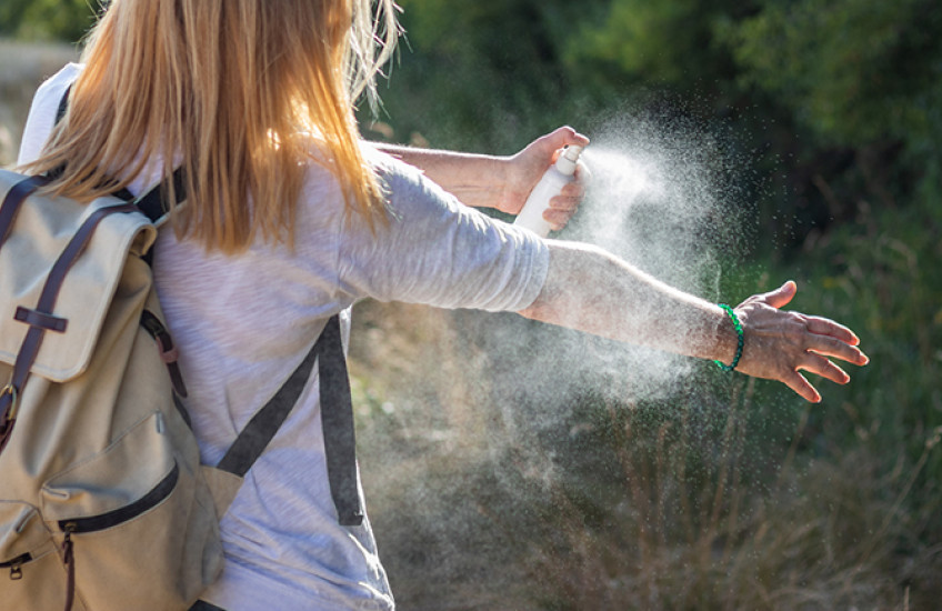 white woman wearing a backpack, viewed from behind, spraying insect repellant onto her bare forearm in a field next to some woods