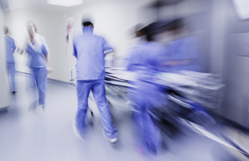 Alt text: Motion blurred image of a group of people in blue hospital scrubs standing, walking, pushing a stretcher around a bend in a brightly lit corridor.