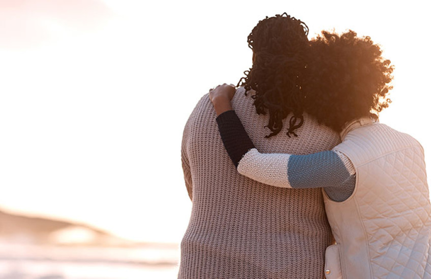 Two women stand side by side embracing one another on a beach facing the ocean.