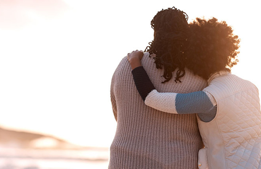 Two women stand side by side embracing one another on a beach facing the ocean.