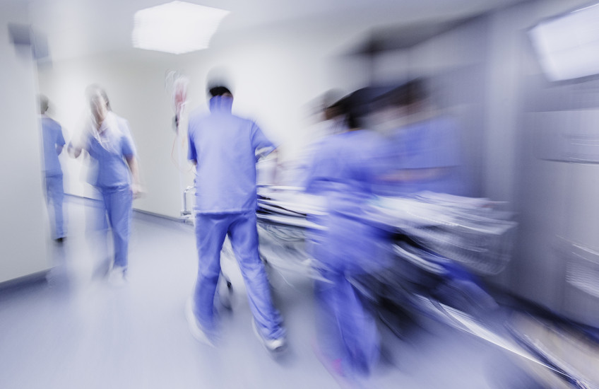 Motion blurred image of a group of people in blue hospital scrubs standing, walking, pushing a stretcher around a bend in a brightly lit corridor.