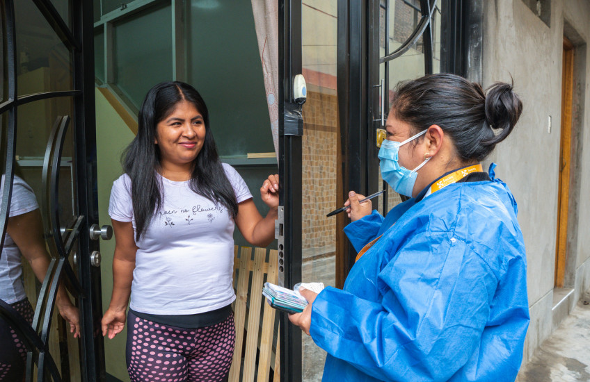 A woman in a t-shirt opens a door to talk with a woman in medical mask and protective robe.