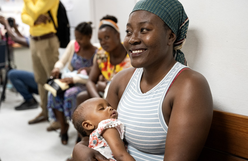 A seated woman in a sleeveless shirt and a headscarf holds a sleeping baby in the foreground, while more people sit in a line behind them in a waiting room. 