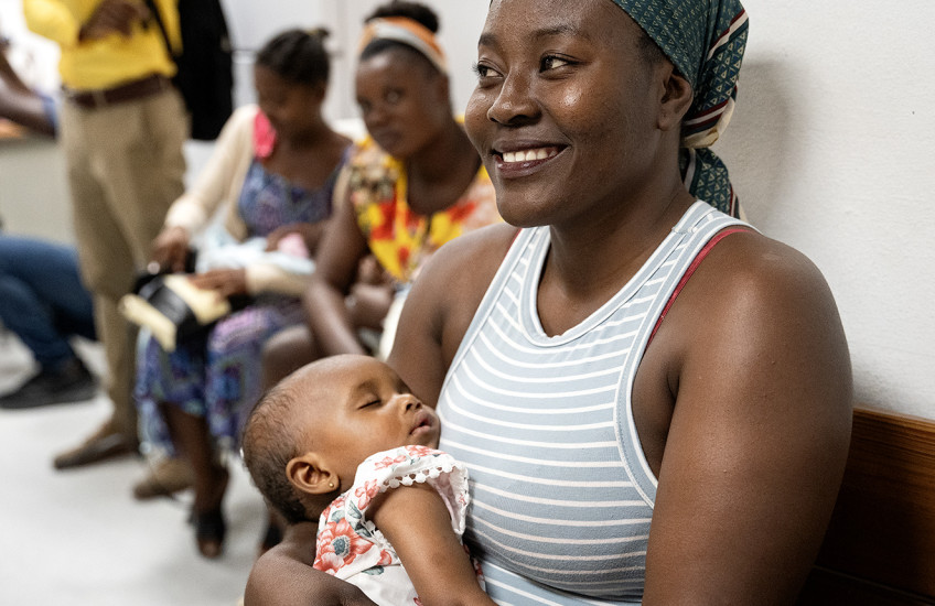 A seated woman in a sleeveless shirt and a headscarf holds a sleeping baby in the foreground, while more people sit in a line behind them in a waiting room. 