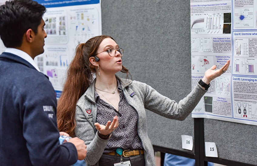 A young woman stands in front of research poster motioning with her hands while a young man listens to her explanation.