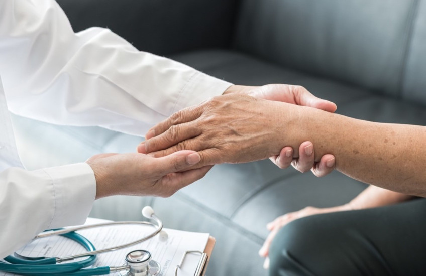 Person in a white coat, presumably a doctor, holds the hand and wrist of a patient. Both people are mostly out of frame. A clipboard and stethoscope rest between them..