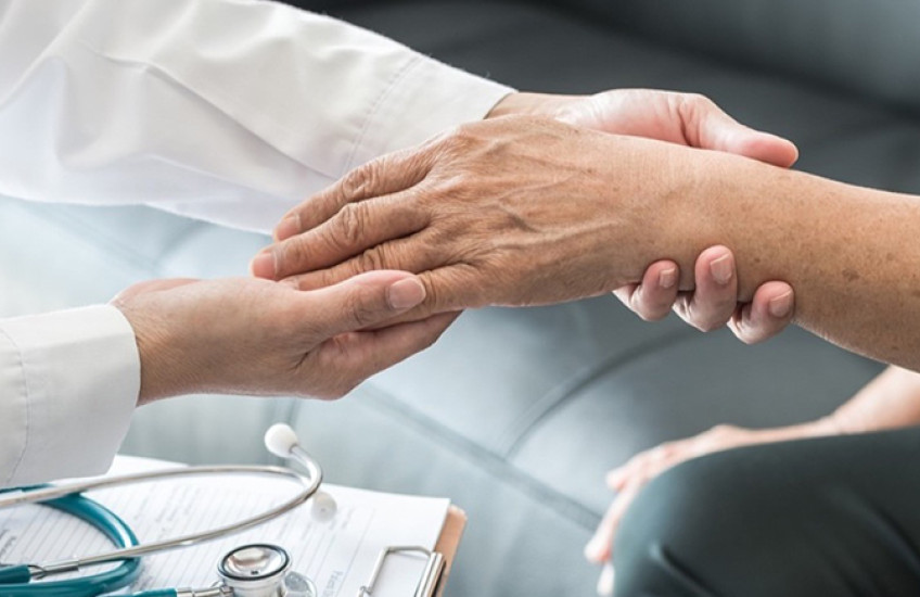 Person in a white coat, presumably a doctor, holds the hand and wrist of a patient. Both people are mostly out of frame. A clipboard and stethoscope rest between them..