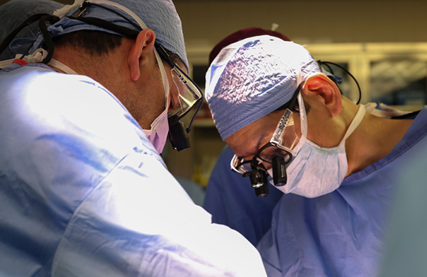Photo of surgeons in scrubs and masks operating on an unseen patient. Only one is seen head-on. He wears attachments on his glasses to magnify what he's seeing.